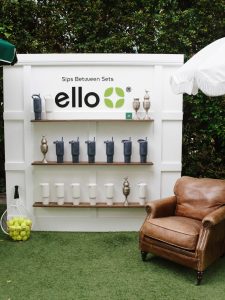 SnapInstato_709770796_18560884081067238_6698127746891094872_n - Lombardi House Outdoor beverage display with wooden shelves holding blue and white tumblers, trophies, and a sign that reads 'ello' with a green leaf logo; tennis ball basket on the left and a brown leather chair on the right, all on artificial grass.