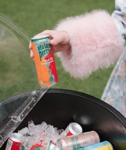 SnapInstato_712429330_18561940876067238_5887612645963327041_n - Lombardi House Person wearing a pink fuzzy sleeve holds an orange-and-green can labeled Maison Perrier French Kiss sparkling juice over a tub filled with ice and other cans.
