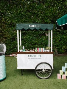 SnapInstato_713744292_18562263535067238_4098529811966525877_n - Lombardi House Green canopy cart labeled 'NAKED LIFE' with non-alcoholic cocktails on a wooden counter, set against a hedge backdrop on artificial grass.